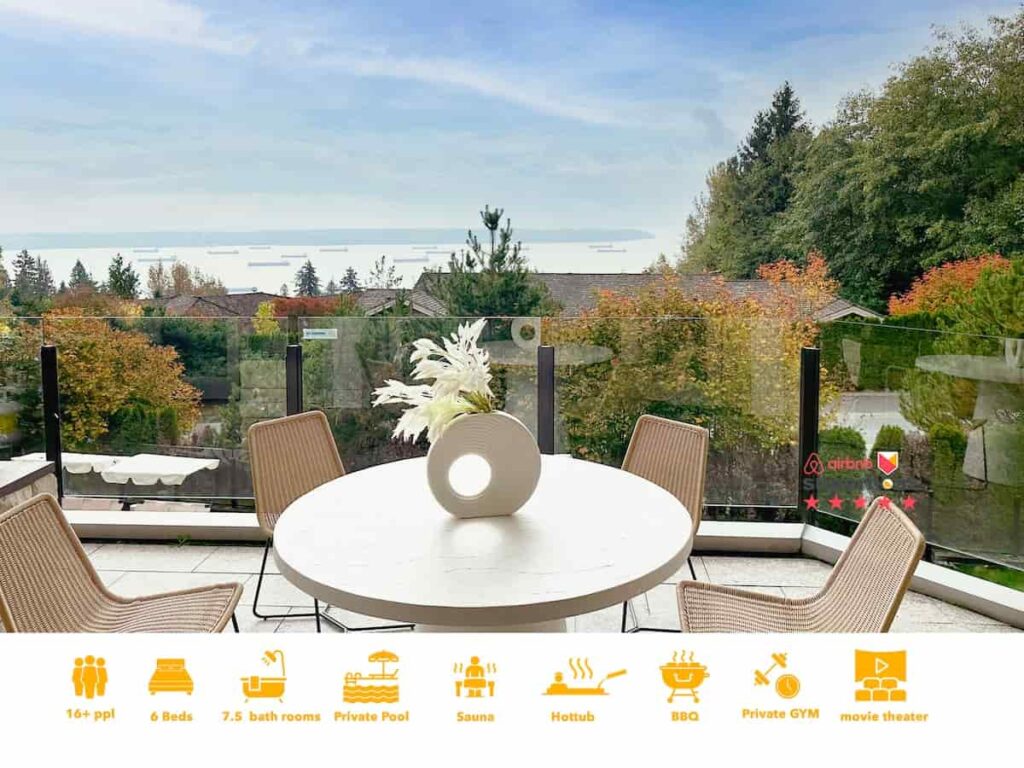 Outdoor patio with wicker chairs around a white circular table, overlooking a serene, tree-filled neighborhood and distant sea view under a blue sky.