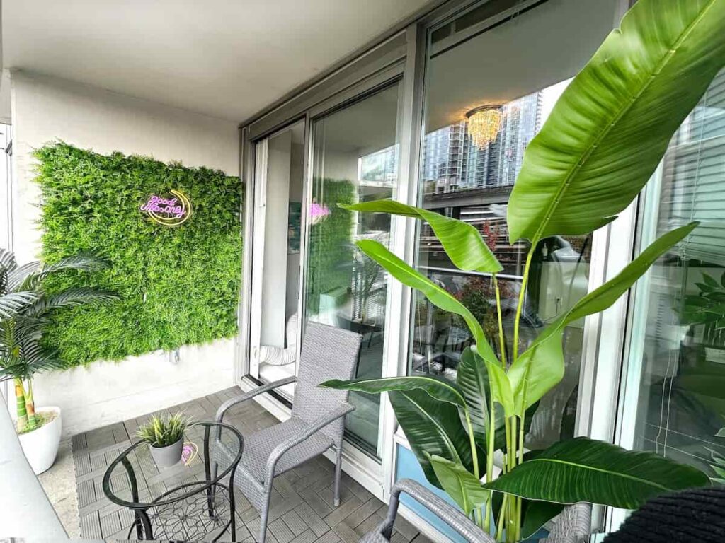 Balcony with glass doors, vibrant green plants, a gray wicker chair, and a small glass table. A neon sign on a foliage wall adds a modern touch. Urban view outside.