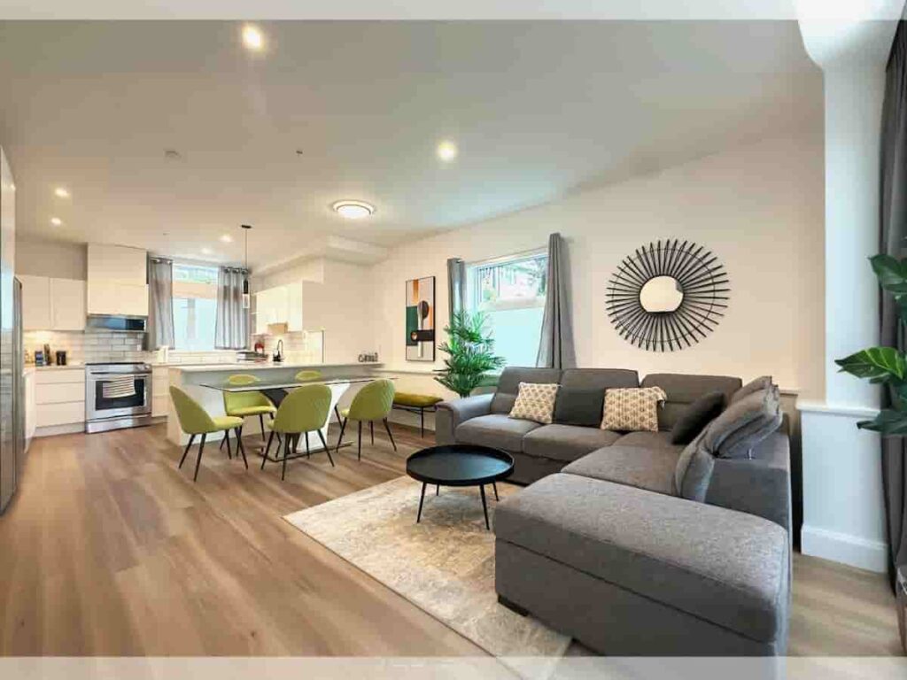 Modern open-plan living room and kitchen with light wood floors. Features gray sectional sofa, green dining chairs, minimalist decor, and bright lighting.