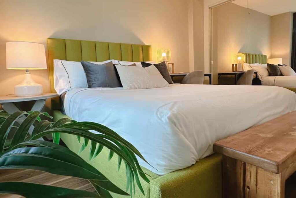 A cozy bedroom with a green upholstered headboard, white bedding, and assorted pillows. Warm lighting, wooden bench, and potted plant add a serene feel.