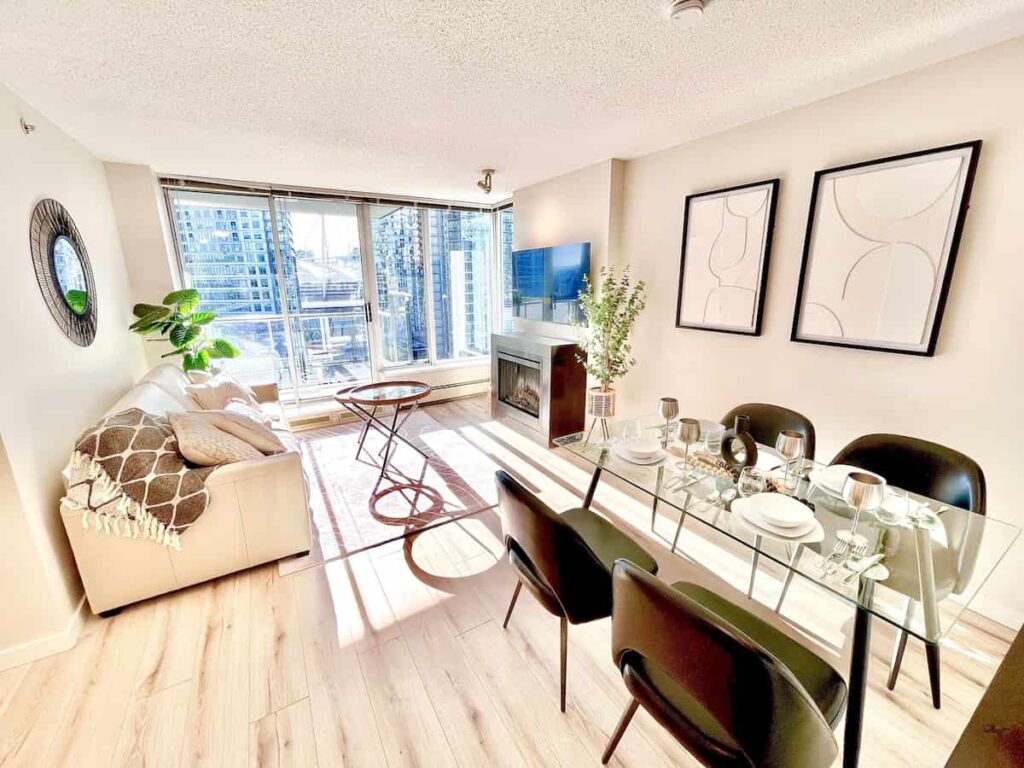 Bright, modern living-dining area with a cream sofa, geometric artwork, glass dining table set, and large windows offering a city view. Cozy and airy.