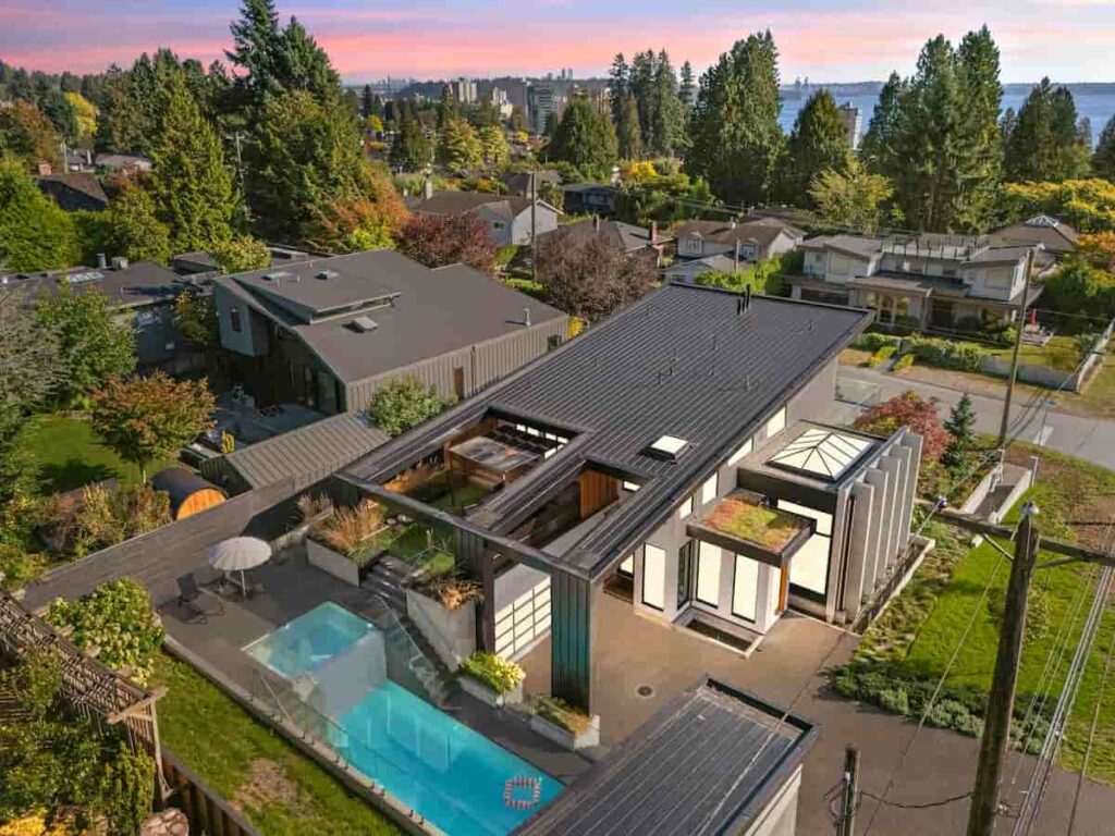 Aerial view of a modern house with a sleek design, featuring a rooftop garden and pool. Surrounded by lush greenery and neighboring homes, under a pink-hued sunset sky.